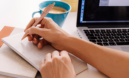 hand writes in a datebook, coffee and laptop nearby