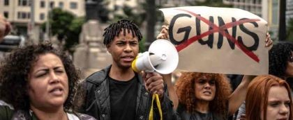 man walking in protest holding a microphone with sign behind stating guns crossed out