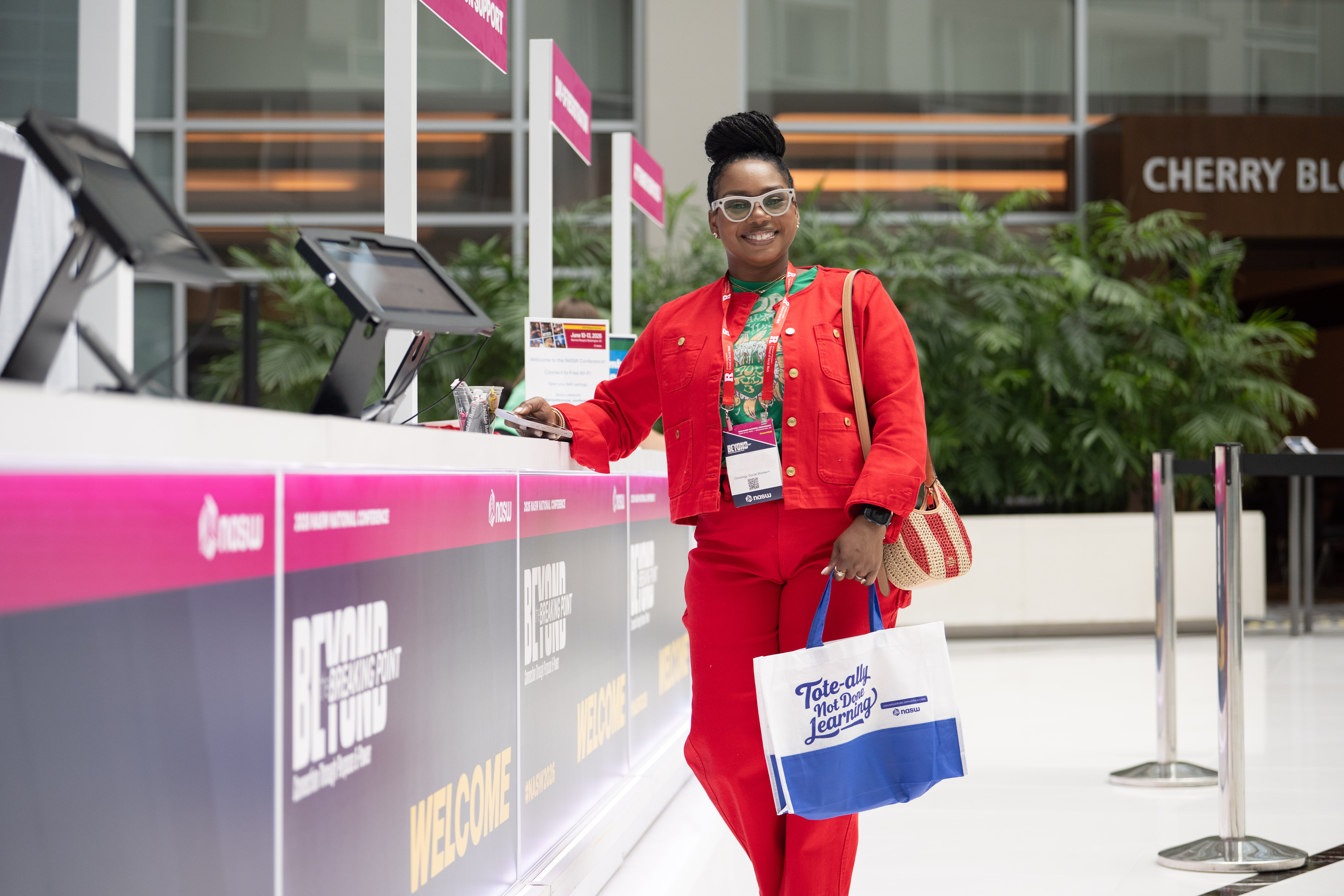 A smiling attendee wearing a red outfit and conference badge stands at the NASW National Conference registration desk while holding an NASW tote bag.