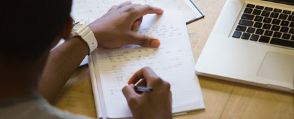 adult writing in notebook next to laptop computer