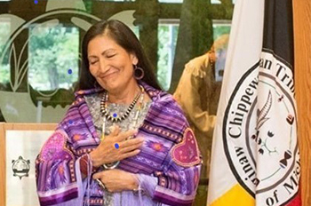 Suzanne Cross smiling with eyes closed, hands over heart, wearing purple quilt with lavender fringe