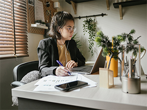 young woman sits at desk with laptop, earbuds, cell phone