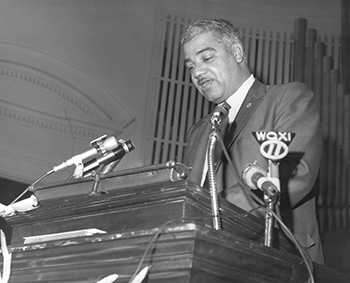 Whitney M. Young Jr speaking at a podium, with broadcast microphones