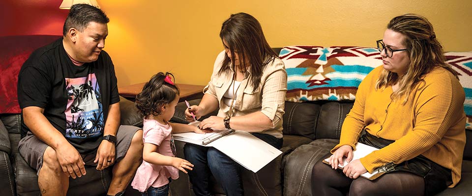 three adults sit on sofas, toddler stands among them