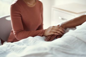 person sitting next to hospital bed holds the hand of a patient