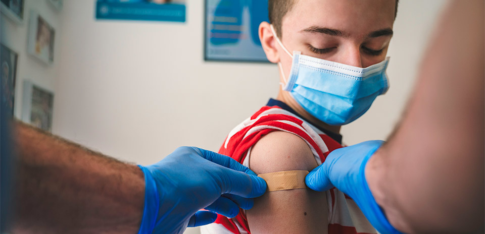 young person wearing face mask getting an adhesive bandage put on their arm by adult wearing surgical gloves