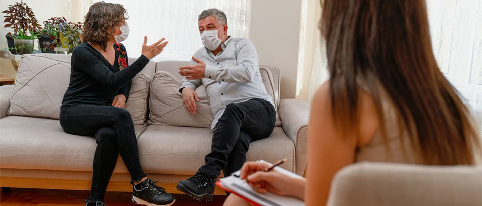 couple wearing face masks talks on sofa while woman with notepad observes