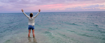 woman stands with her feet in the ocean, arms in the air